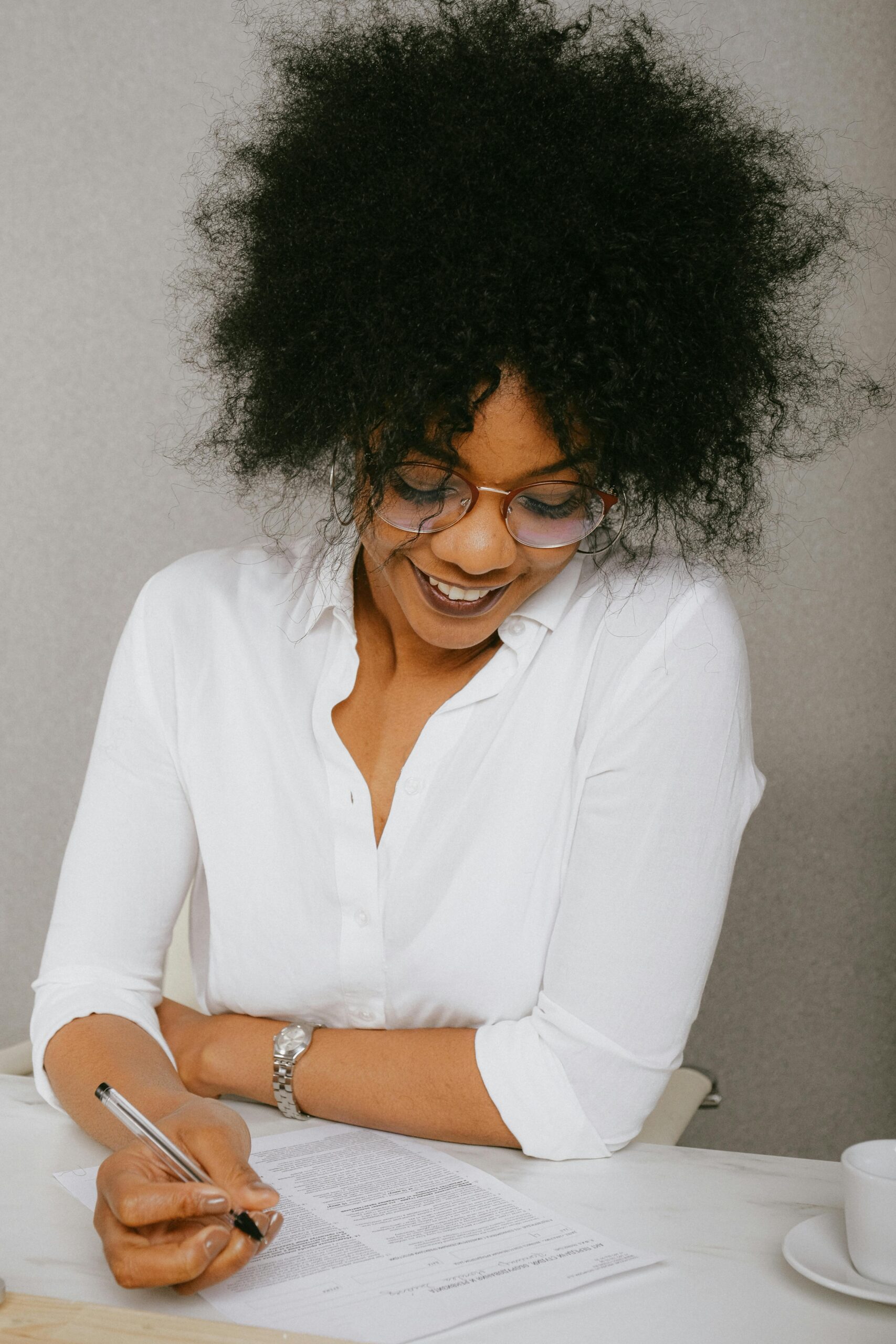 Smiling woman in business attire signing documents at a desk indoors.